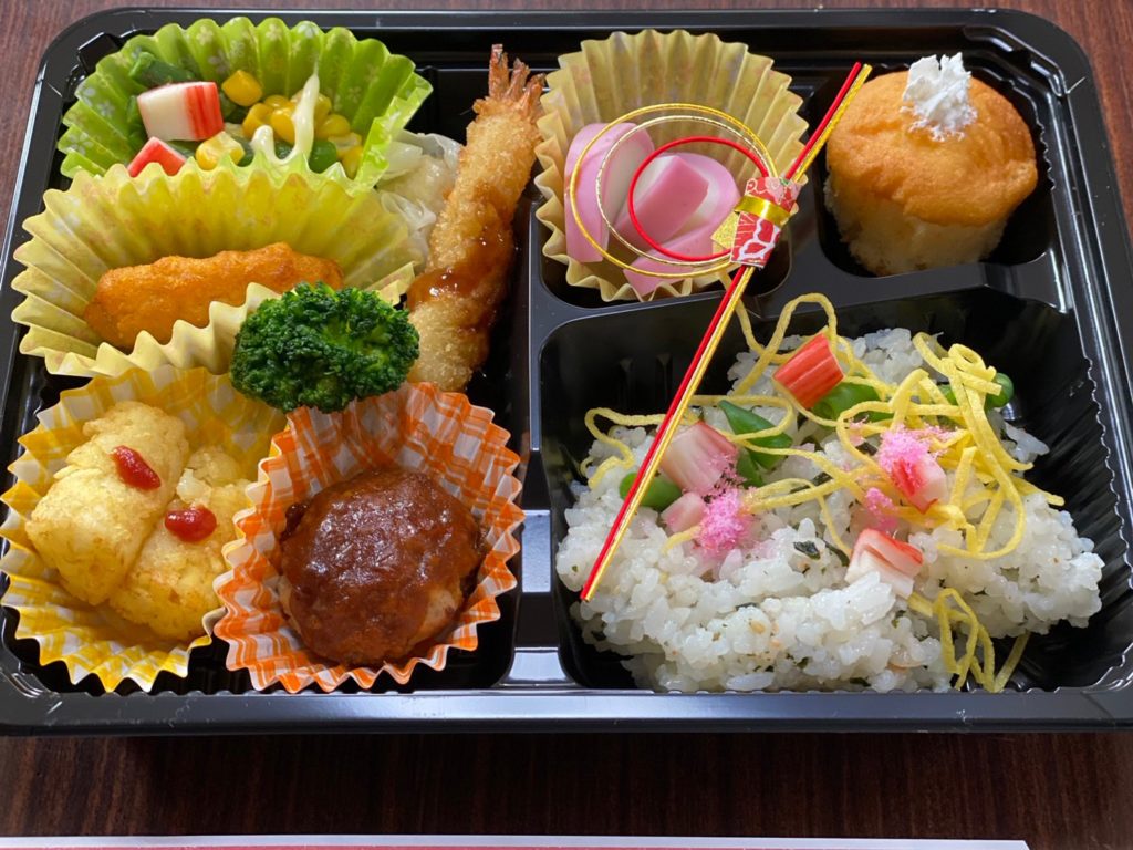 介護施設の食事用弁当。揚げ物や煮込み料理、ご飯が黒いプラスチック容器に彩り良く盛られている。